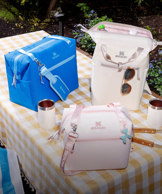 Three Stanley 1913 soft cooler bags in blue, cream, and pink displayed on a gingham-covered outdoor table.