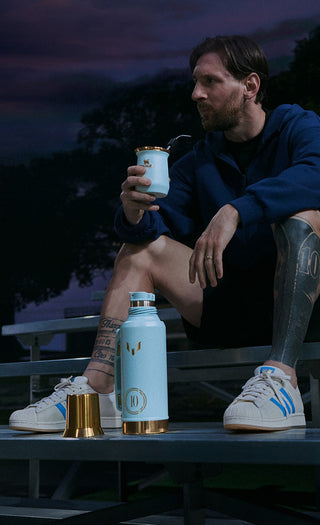 Lionel Messi seated on bleachers holding a Stanley x Messi stainless steel cup, with a matching 1 L bottle and gold cup base at his feet
