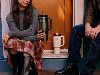 Two people sitting on a porch steps with Stanley 1913 tumblers — dark green and cream — in ribbed style with handles and straws.