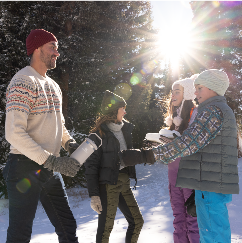 Family enjoying a snowy outdoor day, man holding a white Stanley 1913 insulated thermos, sun shining through pine trees