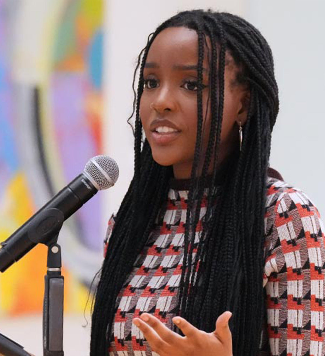 Person speaking at a microphone, wearing a red and black geometric patterned top, with long braided hair.