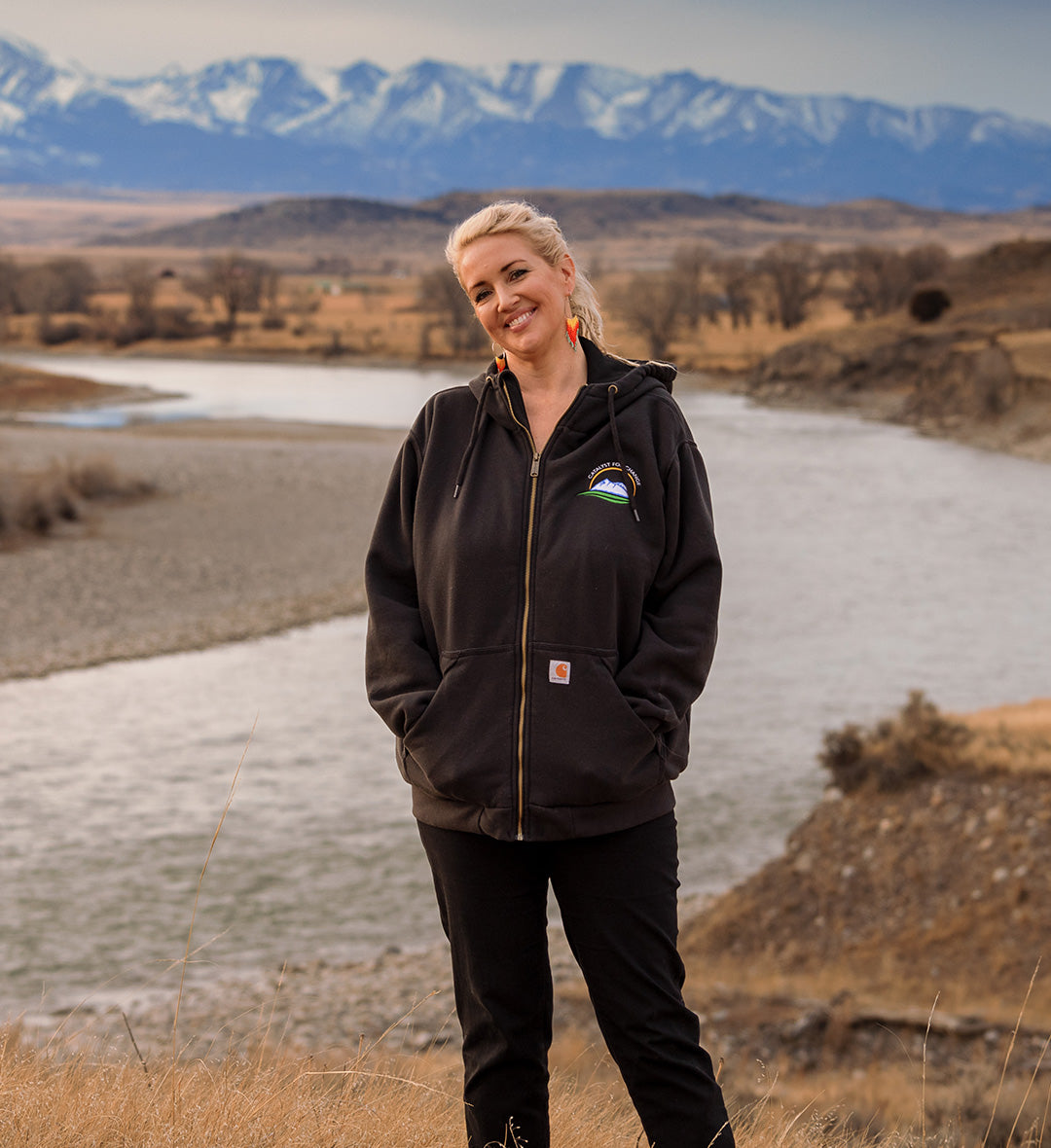 Woman in black Carhartt hoodie smiling outdoors by a river with snow-capped mountains in the background.