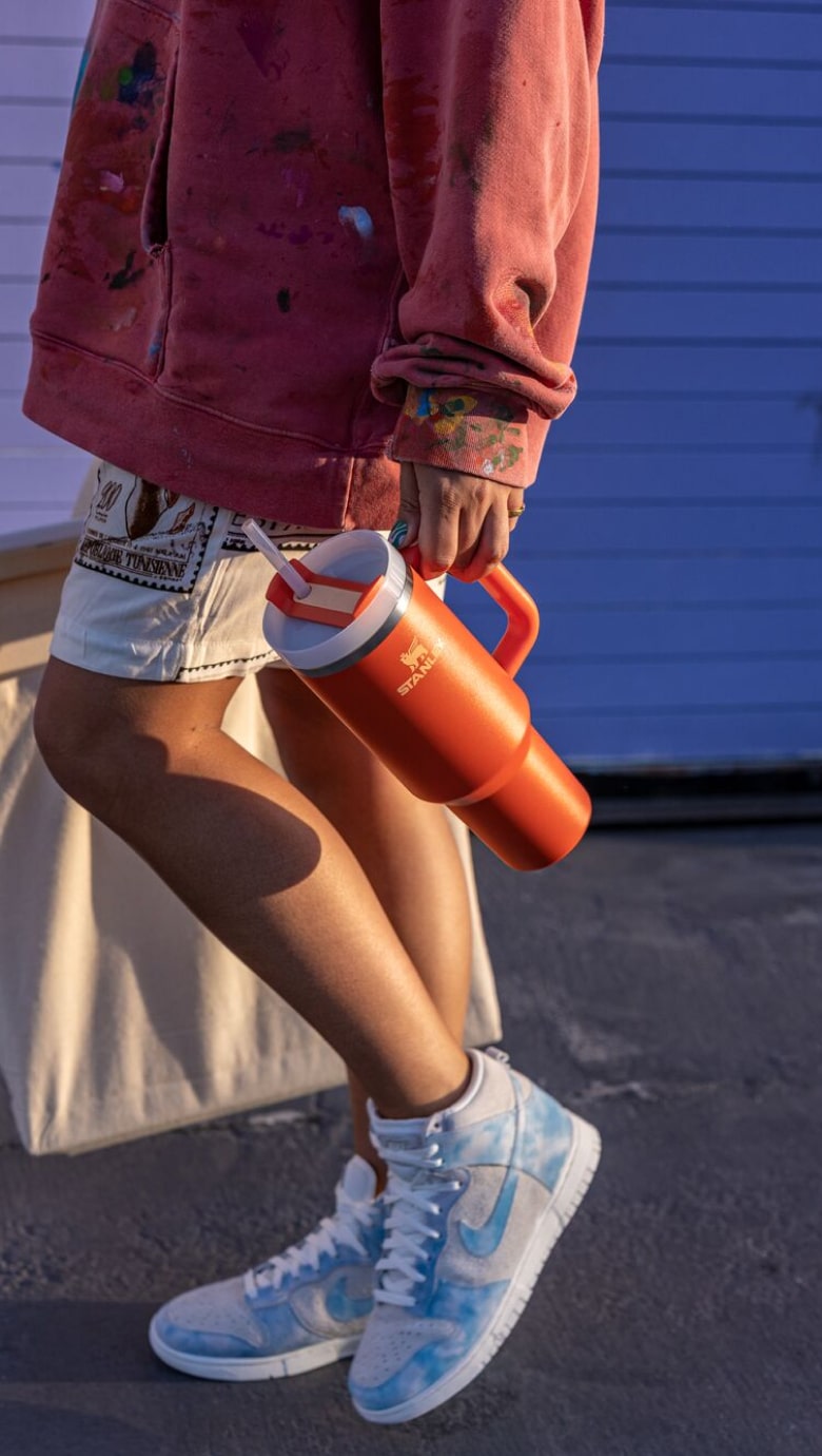 Stanley 1913 orange insulated jug held by person in paint-splattered hoodie, white shorts, and tie-dye Nike Dunks.