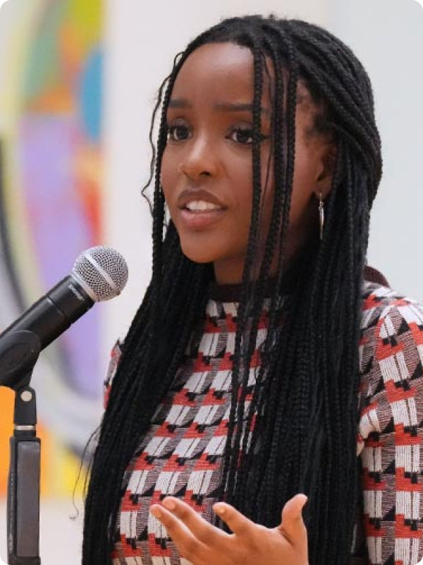 A young woman with long braids speaks at a microphone, gesturing expressively, wearing a red and white patterned top.