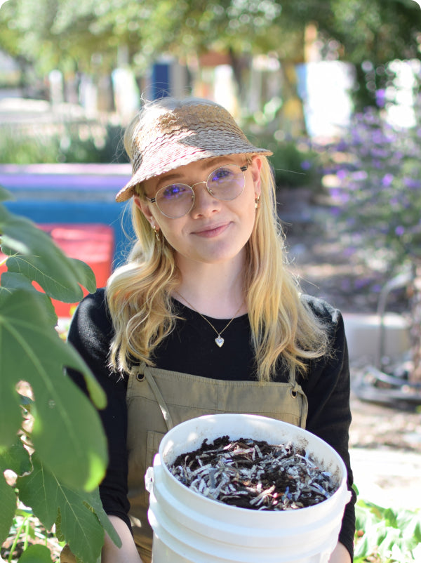 Young woman in straw hat and overalls holding a white bucket of compost in a sunny garden setting.