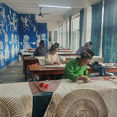 People working at wooden tables in a blue-walled workshop, creating hand-carved relief designs on fabric panels.