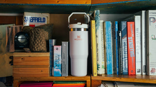 Stanley 1913 white insulated tumbler with handle lid standing upright on a wooden bookshelf surrounded by books